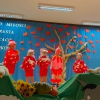 Dzień Babci i Dziadka. Na scenie dziewczynki w czerwonych pelerynkach z białymi piórami i czapkami kurek. W rękach trzymają papierowe jajka. Przed nimi chłopiec przebrany za koguta. W tle kotara z drzewem i sercami oraz napisem. 