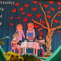 Dzień Babci i Dziadka. Na scenie dzieci w stroju babci i dziadka siedzą przy stoliku. Chłopiec czyta gazetę, babcia ogląda na tablecie. W tle kotara z drzewem, sercami i napisem. Z przodu słoneczniki i dwie dynie.