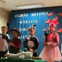 Dzień Babci i Dziadka. Na scenie od lewej dziecko w stroju krowy, dziadka, krowy i świnki. Ostatnia dziewczynka trzyma mikrofon. Z przodu pluszowa gęś. Za nimi kotara z napisem: 