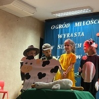 Dzień Babci i Dziadka. Na scenie w rzędzie od lewej stoją dzieci w strojach: dziadka, wnuczka, wnuczki i krowy. Ostatnia dziewczynka trzyma mikrofon. Przed nimi krowa z papieru. W tle kotara z napisem: 