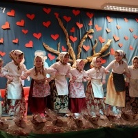 Dzień Babci i Dziadka. Osiem dziewczyn w długich spódnicach i fartuszkach, białych bluzkach z chustami na głowie stoi trzymając się pod ręce. Z przodu prezenty zapakowane w celofan i ozdobione papierowymi sercami. W tle kotara z drzewem i sercami.
