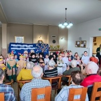 Dzieci u Seniorów. Seniorzy siedzą na krzesłach, patrzą na dzieci. Dzieci w strojach z różnych stron świata śpiewają seniorom piosenkę. W tle tablica.
