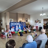 Dzieci u Seniorów. Dziewczynki w ludowych strojach siedzą na krzesłach. W rękach mają gazetę. Po lewej stronie przy stoliku chłopiec i dziewczynka w strojach dziadków. Na dzieci patrzą seniorzy.