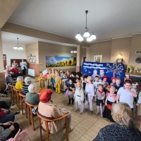 Dzieci u Seniorów. Dzieci przebrane w stroje z różnych stron świata stoją przed seniorami. Śpiewają piosenkę. Za nimi tablica.