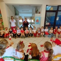 Pokaz Mody Patriotycznej. Dzieci w biało-czerwonych ubiorach siedzą na ławeczkach w rzędzie. Za nimi dwie panie w strojach ludowych. Z drugiej strony dzieci na krzesłach patrzą w ich stronę.  