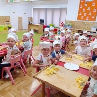 Sałatka owocowa. Dzieci siedzą przy stolikach ubrane w kuchenne fartuszki i czapki kucharskie. Kroją owoce do sałatki.