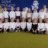 Dzień Edukacji Narodowej. Dzieci ustawione w dwóch rzędach pozują do zdjęcia ma tle tablicy z wizerunkiem nauczycielki i napisem Dzień Edukacji Narodowej.