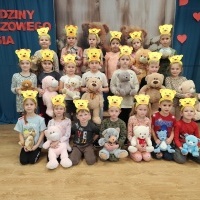 Dzień Misia. Dzieci ustawione w trzy szeregi pozują do zdjęcia. Na głowach mają opaski z twarzami misia a w rękach trzymają pluszowe maskotki. W tle kotara z napisem: Urodziny pluszowego misia.