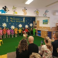 Koncert wiosenny. W sali zajęć rodzice patrzą w kierunku dzieci ubranych w stroje zwierząt łąkowych. Dziewczynka w stroju pani wiosny mówi do mikrofonu.