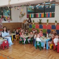 Śniadanie Wielkanocne. Dzieci elegancko ubrane siedzą przy świątecznych stołach i pozują do zdjęcia. Przy osobnym stole siedzą 4 panie.