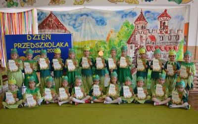 Dzieci z Grupy Żabki ustawione jedno obok drugiego, w strojach żabek w kolorze zielonym. W tle tablica z napisem Dzień Przedszkolaka - wrzesień 2020 i malowany na płótnie zamek. 