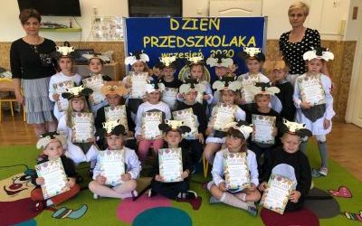 Dzieci z Grupy Kotki. Ustawione jedno obok drugiego. Dzieci w strojach biało, szaro czarnych. W tle tablica z napisem: 